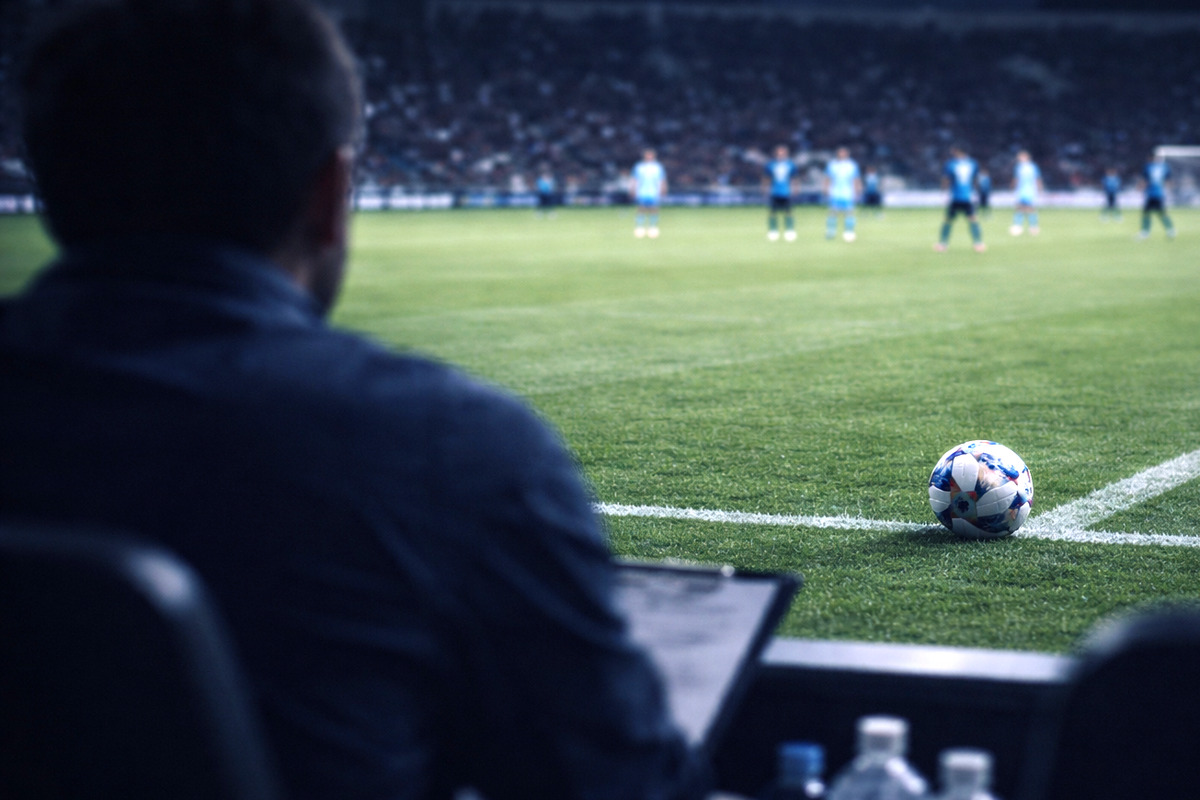 ベンチからピッチを見つめる視点。手前にスタッフのシルエットとメモ、ライン際のボールと遠くで展開するプレー