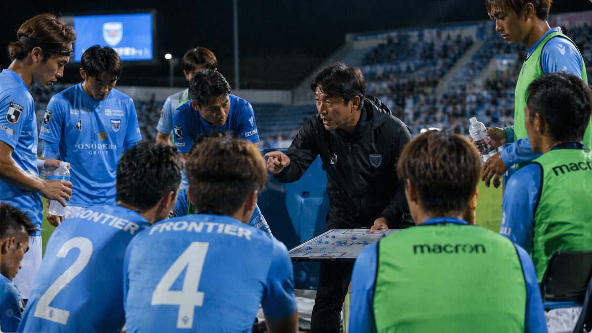 ハーフタイムにベンチ前で監督が選手に戦術を伝えている静かなシーン